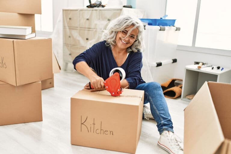 Middle Age Grey Haired Woman Smiling Happy Packing Kitchen Cardboard Box At New Home.
